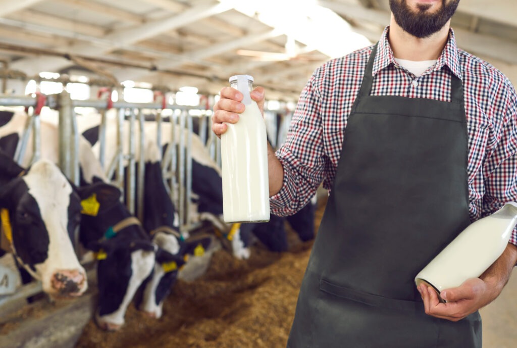 Cropped male farmer holding glass bottles of fresh organic milk with cows in background