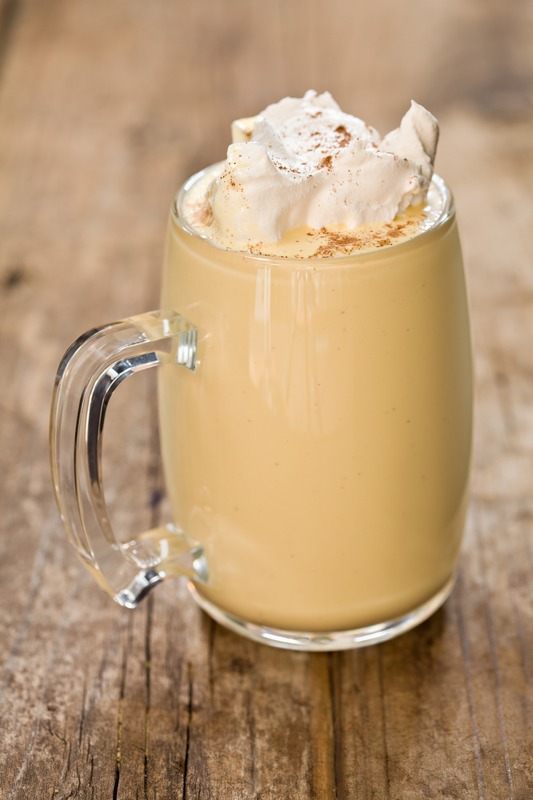 A close up vertical shot of a clear mug full of eggnog topped with whipped cream and ground nutmeg. Shot on a grungy old wooden table.